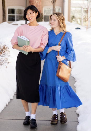 2 women walking outdoors on the sidewalk holding a book bag and books